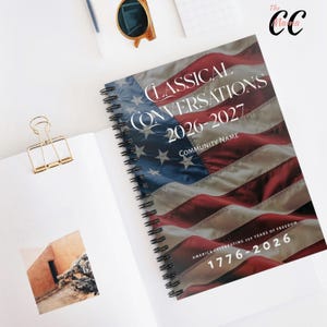 May include: A spiral-bound notebook with a cover featuring the American flag design. The cover reads "Classical Conversations 2026-2027" and "America Celebrating 250 Years of Freedom 1776-2026". The notebook is open on a white surface.