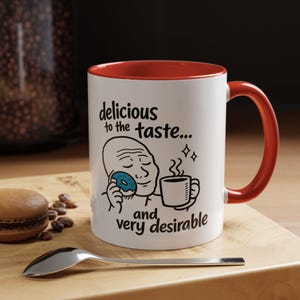 May include: White ceramic mug with a red interior and handle. The mug features a cartoon illustration of a person eating a donut and drinking coffee, with the text "delicious to the taste... and very desirable."