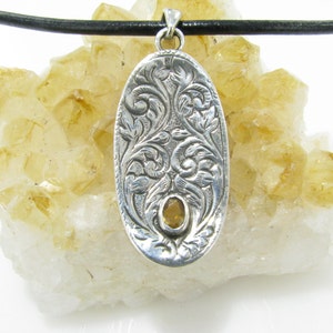 May include: A silver oval pendant with an intricate floral design and a yellow gemstone in the center. The pendant is hanging from a black leather cord.
