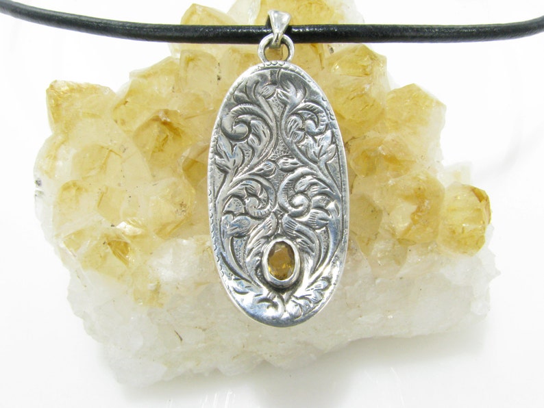 May include: A silver oval pendant with an intricate floral design and a yellow gemstone in the center. The pendant is hanging from a black leather cord.