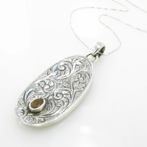 May include: A silver pendant necklace with an oval-shaped charm featuring an intricate floral design. The charm has a small, round, yellow gemstone in the center.