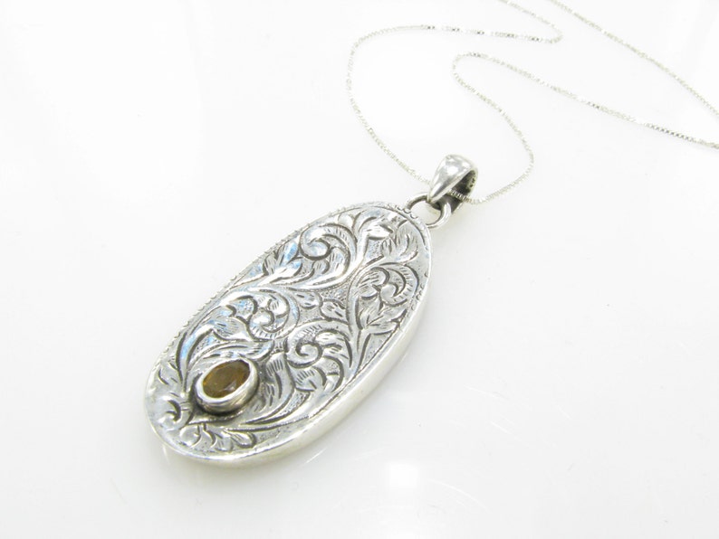 May include: A silver pendant necklace with an oval-shaped charm featuring an intricate floral design. The charm has a small, round, yellow gemstone in the center.