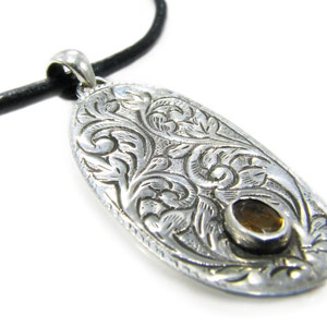 May include: A silver pendant with an intricate floral design and a small, round, amber-colored stone in the center. The pendant is attached to a black leather cord.