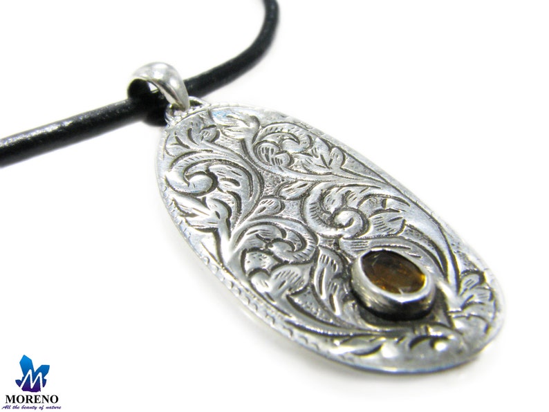 May include: A silver pendant with an intricate floral design and a small, round, amber-colored stone in the center. The pendant is attached to a black leather cord.