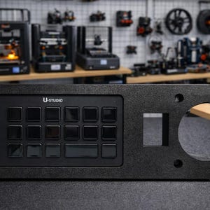 May include: A black control panel with the text "U-STUDIO" and multiple square buttons. The panel has a rectangular cutout and two circular holes. The background shows 3D printers and other equipment.