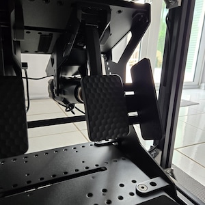 May include: Black racing simulator pedal set with textured foot pads. The pedals are mounted on a black metal frame with various mounting points and adjustable components. The image shows the underside of the simulator.