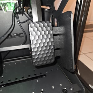 May include: Close-up of a black racing simulator pedal with a textured, honeycomb-like surface. The pedal is mounted on a black metal frame, with visible screws and other components. The image shows a part of a racing simulator setup.