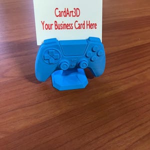 Könnte beinhalten: Ein hellblauer 3D-gedruckter Visitenkartenhalter in Form eines Videospiel-Controllers. Er hält eine weiße Karte mit rotem Text: "CardArt3D Your Business Card Here". Der Controller steht auf einer Holzoberfläche.