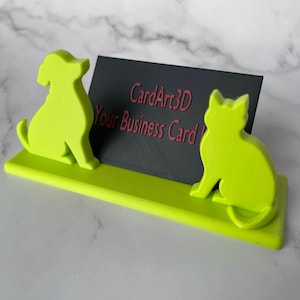 Puede incluir: Un tarjetero verde neón con diseños de siluetas de perro y gato. El soporte tiene una base rectangular y una tarjeta gris oscuro con el texto "CardArt3D Your Business Card".