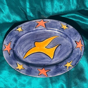 May include: Oval ceramic serving platter with a dark blue background. The platter features an orange bird in the center and is surrounded by orange and yellow stars. The hand-painted design adds a unique touch to this decorative piece.