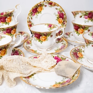 May include: A set of white porcelain teacups and saucers with a floral rose pattern in shades of red, pink, and yellow. The teacups have gold trim and handles. A lace glove and a tea bag are also present.