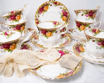 May include: A set of white porcelain teacups and saucers with a floral rose pattern in shades of red, pink, and yellow. The teacups have gold trim and handles. A lace glove and a tea bag are also present.