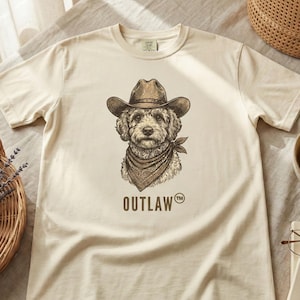 Puede incluir: Camiseta color crema con una ilustración detallada de un perro con sombrero de vaquero y pañuelo. La palabra "OUTLAW" está impresa debajo del perro. La camiseta está tendida sobre una superficie neutra.