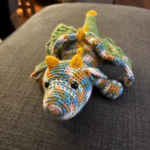Può includere: Un drago all'uncinetto fatto a mano nei toni del blu, arancione, verde e giallo. Il drago ha piccoli occhi neri, corna gialle e ali verdi. È appoggiato su una superficie grigia.