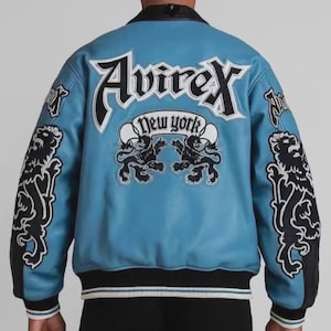 May include: A light blue leather jacket with black and white detailing. The back displays the word "Avirex" in large white letters, with "New York" underneath. Lion graphics adorn the sleeves and back. The jacket has a black collar and cuffs.