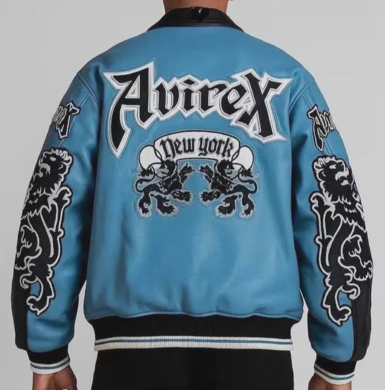 May include: A light blue leather jacket with black and white detailing. The back displays the word "Avirex" in large white letters, with "New York" underneath. Lion graphics adorn the sleeves and back. The jacket has a black collar and cuffs.