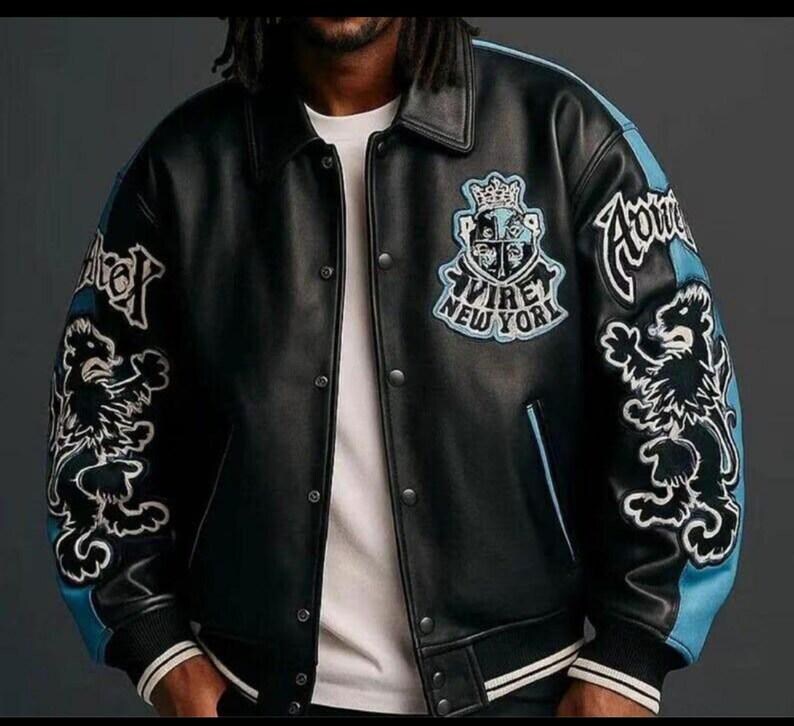May include: Black leather jacket with light blue detailing on the sleeves and collar. It features embroidered lion graphics and the text "AVIREY NEW YORK". The jacket has a button-up front and a ribbed hem.