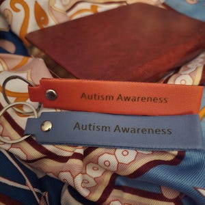 May include: Two leather keychains, one orange and one blue, with the text "Autism Awareness" printed on them. The keychains are resting on a patterned fabric with a brown leather-bound book in the background.
