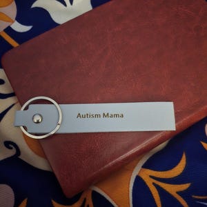 May include: A light blue leather keychain with the text "Autism Mama" is placed on a burgundy leather notebook. The background features a patterned fabric with blue, white, and orange floral designs.