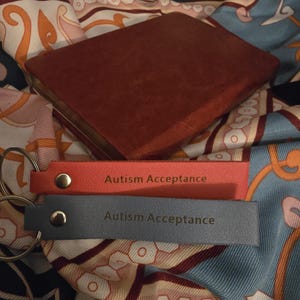 May include: A red leather-like keychain and a gray leather-like keychain with the words "Autism Acceptance" engraved on them. A brown leather-bound book is in the background. The items are on a patterned fabric.