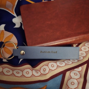 May include: A light blue leather keychain with the text "Autism Dad" in gold lettering. The keychain is resting on a patterned scarf and next to a brown leather notebook.