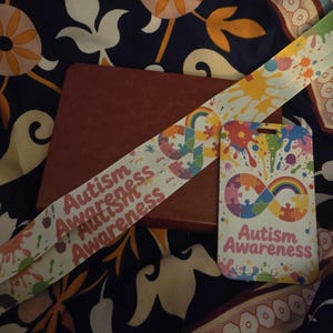 May include: A red leather-bound notebook with a white lanyard and a matching card featuring the words "Autism Awareness." The lanyard and card have colorful paint splatters and a rainbow puzzle piece design.