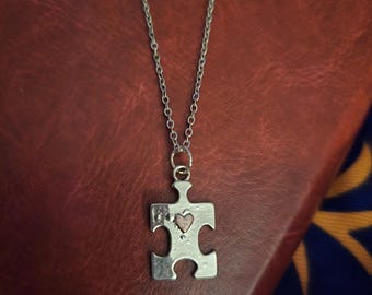 May include: Silver-toned puzzle piece pendant necklace with a small heart cutout. The pendant hangs from a delicate chain. The necklace is displayed on a textured, reddish-brown surface.