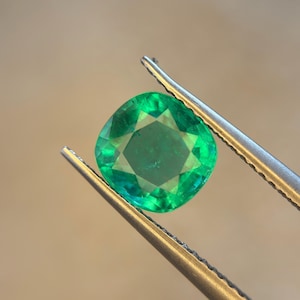 May include: A vibrant, cushion-cut emerald gemstone with a brilliant green hue and multifaceted surface, held securely by a pair of silver-toned metal tweezers. The gemstone displays internal clarity and light reflection across its polished facets.