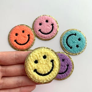 Peut inclure: Plusieurs écussons smiley colorés. Chaque écusson présente une couleur différente, notamment orange, rose, jaune, bleu et violet, avec un sourire noir brodé. Les écussons ont une bordure dorée scintillante.