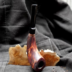 May include: A handcrafted tobacco pipe with a curved, reddish-brown bowl and a black stem. The pipe features a copper accent ring and rests on a piece of light-colored wood. The background is a dark, textured fabric.