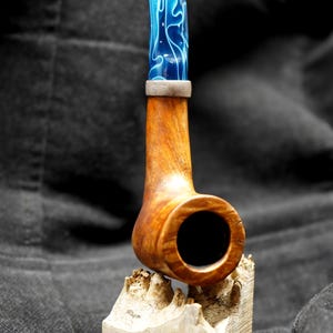May include: A handcrafted tobacco pipe with a wooden bowl and stem. The stem has a blue and white swirl design. The pipe rests on a small, carved wooden base. The pipe is a light brown color.
