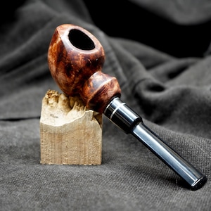 May include: A handcrafted tobacco pipe with a dark brown, polished bowl and a black stem. The pipe rests on a small, rough-hewn wooden block. The bowl has a rich, wood-grain pattern. The stem is accented with silver-colored metal rings.