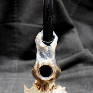 May include: A handcrafted tobacco pipe with a black stem and a uniquely carved wooden bowl. The bowl has a dark, textured finish and is mounted on a light-colored wooden base. The pipe is designed for smoking.