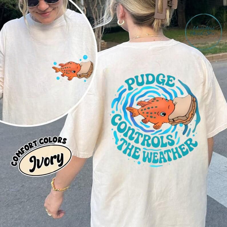A person wearing a white t-shirt with a graphic design of an orange fish and the text "Pudge Controls the Weather" printed on it.