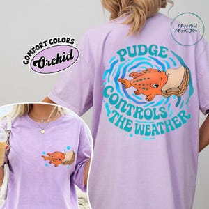 A person wearing a purple t-shirt with a graphic design featuring a fish and the text "Pudge Controls the Weather".