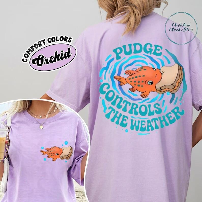A person wearing a purple t-shirt with a graphic design featuring a fish and the text "Pudge Controls the Weather".