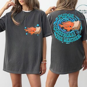 two different views of a woman wearing a gray t-shirt with a graphic design on the back. The design features an orange fish and the text "Pudge Controls the Weather".