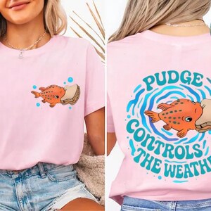 A woman wearing a pink t-shirt with a cartoon fish and the text "Pudge Controls the Weather" printed on it.