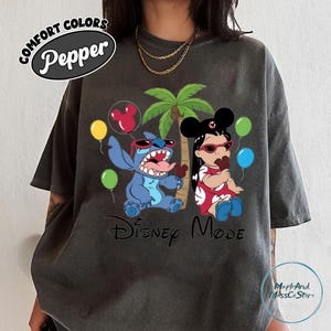 A person wearing a black t-shirt with a graphic design featuring two Disney characters, Stitch and Minnie Mouse, along with various Disney-themed elements such as balloons and a palm tree.