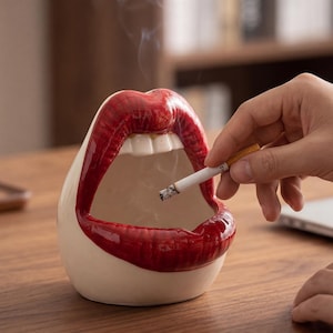 Puede incluir: Un cenicero de cerámica con forma de labios rojos con dientes blancos. El cenicero es blanco con labios rojos y sostiene un cigarrillo encendido. El humo se eleva del cigarrillo.