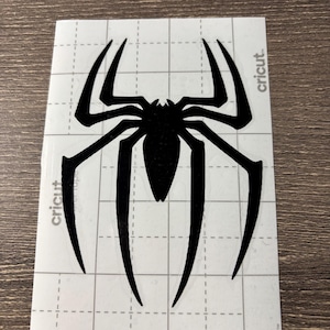 May include: Black spider decal with eight legs and a segmented body. The decal is on a white, grid-patterned backing with the word "cricut" printed on the side.
