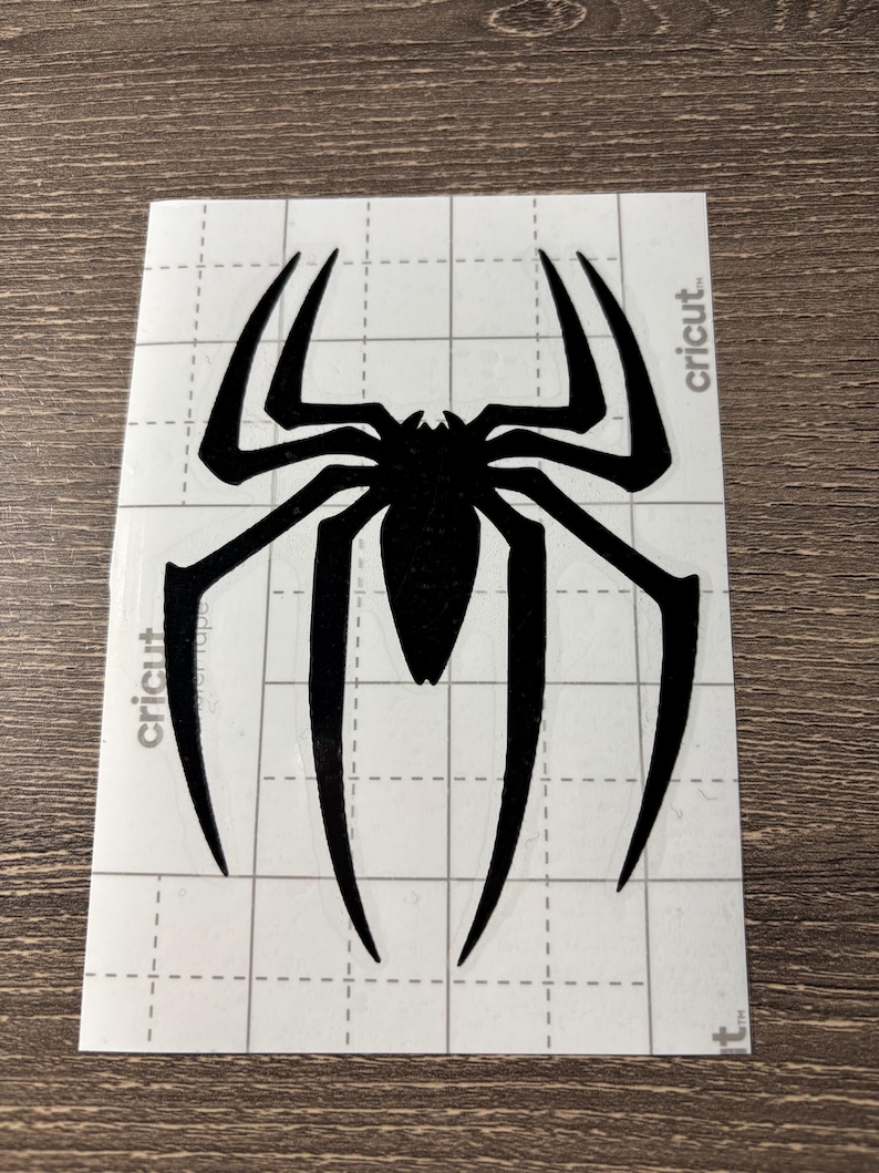May include: Black spider decal with eight legs and a segmented body. The decal is on a white, grid-patterned backing with the word "cricut" printed on the side.
