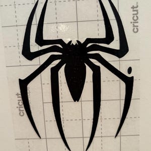 May include: A black spider decal featuring eight elongated, pointed legs and a detailed body. The decal is on a clear backing with a grid pattern, and the word "cricut" is visible. Ideal for crafting and DIY projects.