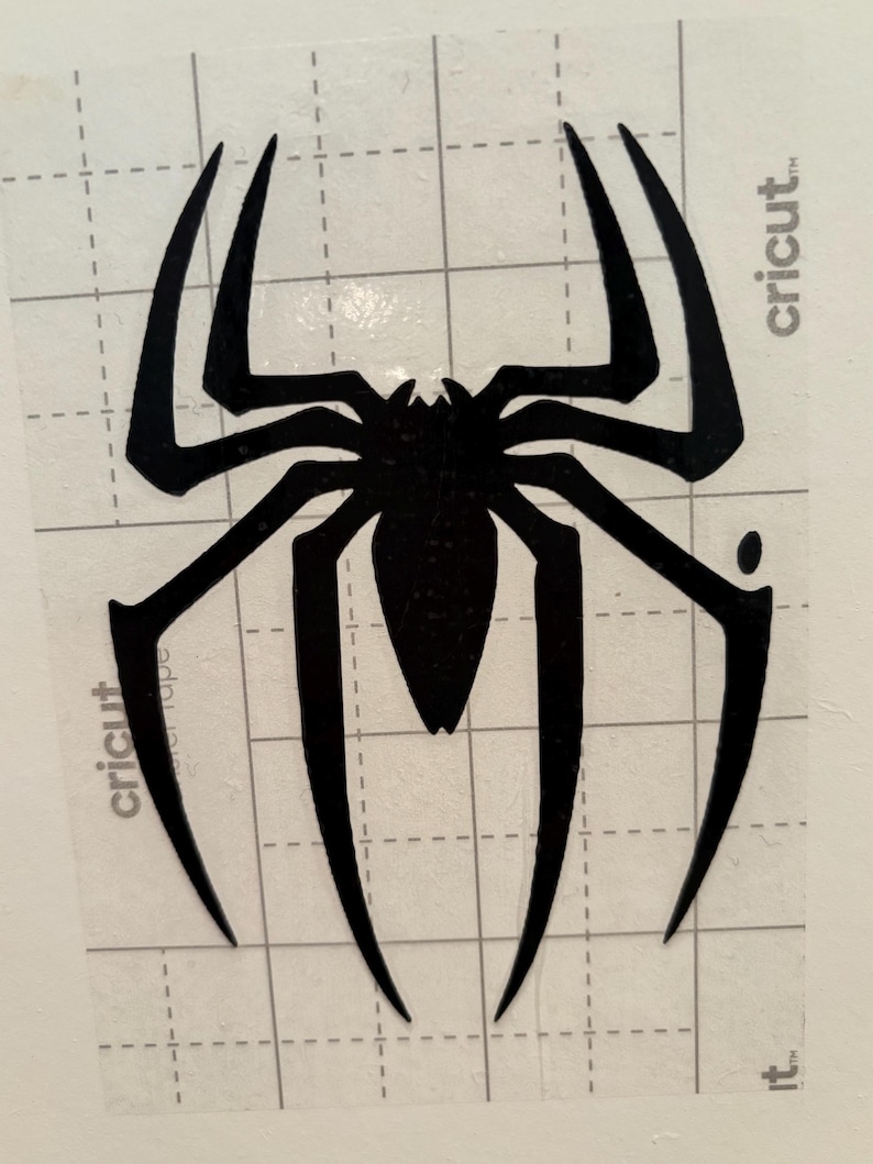 May include: A black spider decal featuring eight elongated, pointed legs and a detailed body. The decal is on a clear backing with a grid pattern, and the word "cricut" is visible. Ideal for crafting and DIY projects.