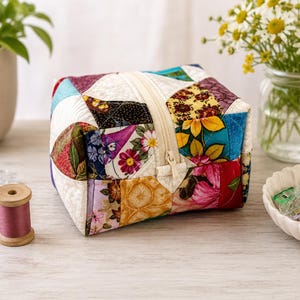 Könnte beinhalten: Ein quadratisches Reißverschlussetui aus einem Patchwork aus gesteppten Stoffquadraten mit verschiedenen Blumen- und geometrischen Mustern in Rot-, Gelb-, Blau-, Lila- und Weißtönen, mit einem cremefarbenen Reißverschluss in der Mitte.
