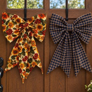 May include: Two large decorative fabric bows hanging on a wooden door. The left bow features a vibrant sunflower pattern with yellow, orange, and red flowers on a cream background. The right bow has a dark blue and tan plaid checkered pattern.