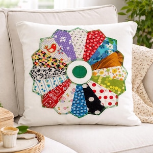 May include: A white square pillow with a colorful, circular patchwork design. The patchwork features various fabric patterns, including floral, polka dots, and geometric shapes, arranged around a green center. A small red bird is stitched on the patchwork.
