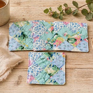 May include: A set of three quilted fabric coasters and one square coaster. The coasters feature a patchwork design with floral patterns in shades of blue, green, and pink. The coasters are arranged on a light wood surface.