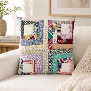 May include: Square patchwork pillow with a variety of floral and geometric patterns in shades of blue, red, purple, and white. The pillow is made of quilted fabric and has a white square in the center of each section. The pillow is on a white couch.