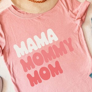 May include: A light pink t-shirt with the words "MAMA MOMMY MOM" in white and pink bubble letters. The shirt is folded, with a straw hat and blue jeans visible in the background. White sneakers are also in the image.